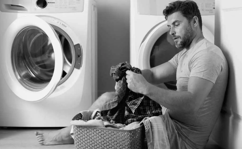 A person sitting beside a washing machine which is not spinning with a bunch of cloths